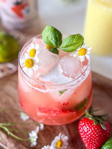 A refreshing strawberry ginger mocktail garnished with basil and chamomile flowers, served on a wooden tray surrounded by chamomile flowers with strawberries, a mason jar full of ginger juice and a lime nearby.